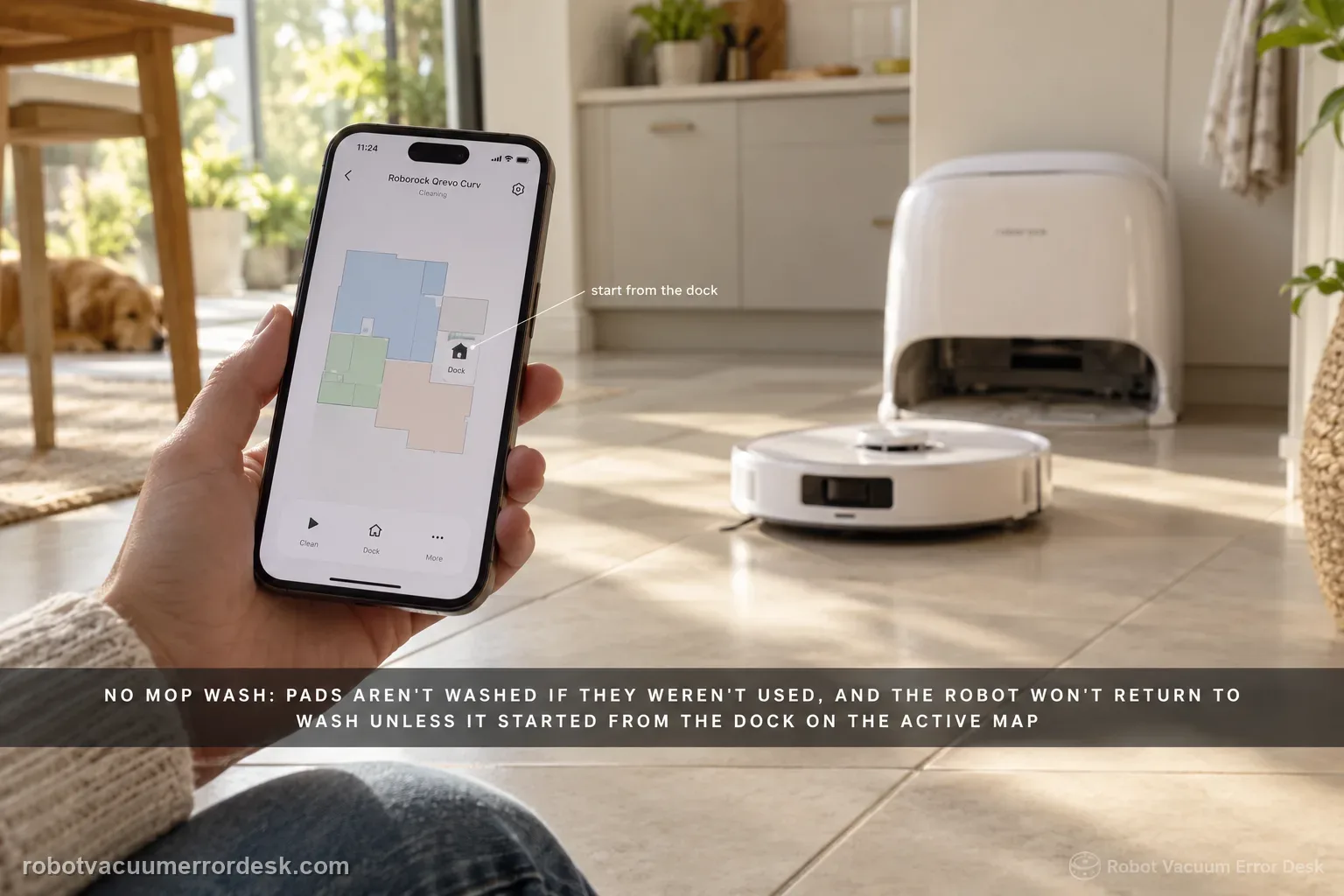 A Roborock Qrevo Curv heading back toward its dock across a kitchen floor, a hand in the foreground holding a phone showing the app map with the dock marked.